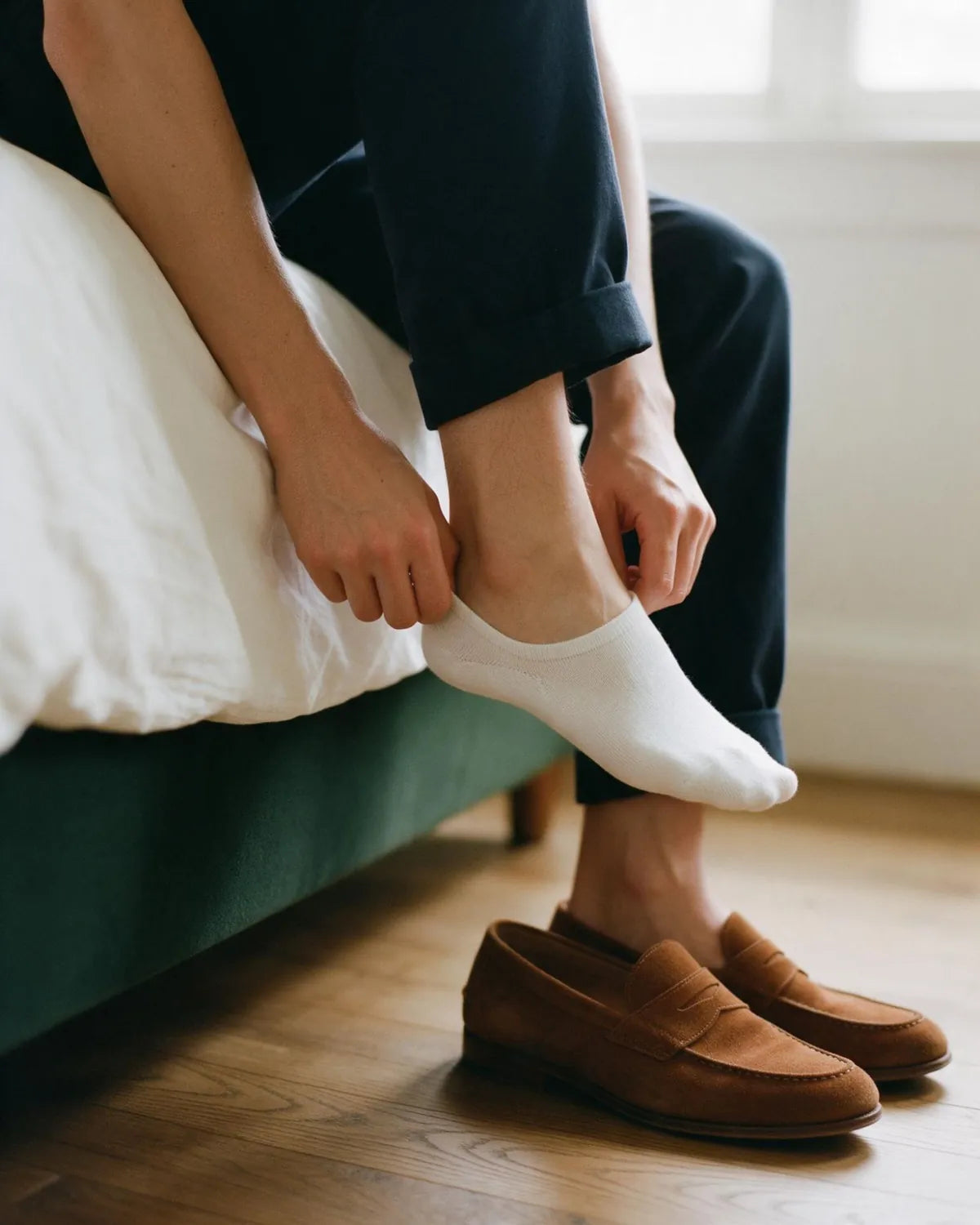 Someone on the edge of a bed easing on a white no-show sock beside a pair of tan suede loafers