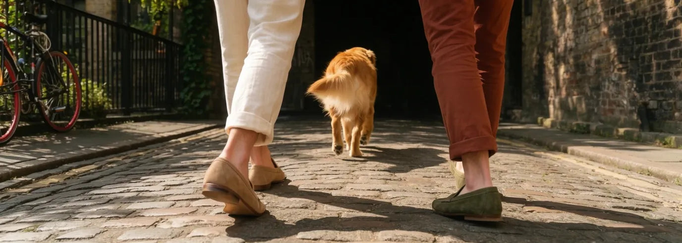 Two people in suede loafers walking down a sunny cobbled lane behind a golden retriever