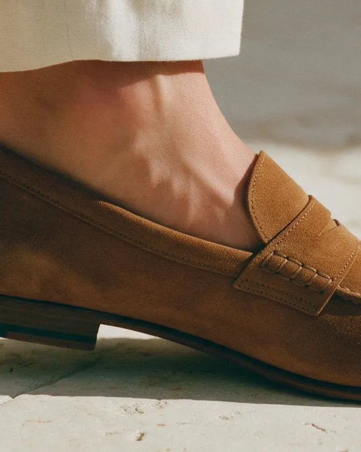 A bare ankle above a tan suede loafer on a pale stone floor