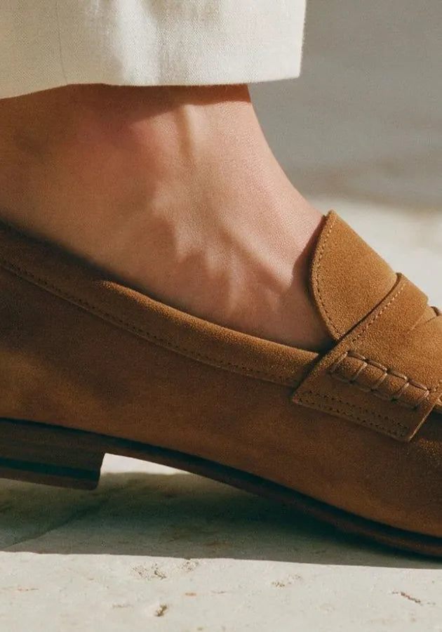 A bare ankle above a tan suede loafer on a pale stone floor