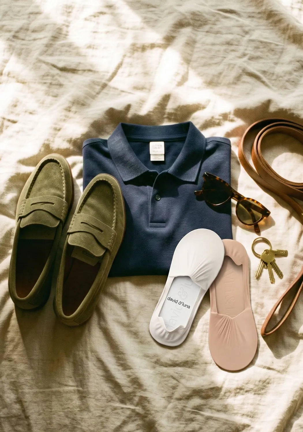 Olive suede loafers, a folded navy polo and no-show socks laid out on linen with sunglasses, keys and a dog lead